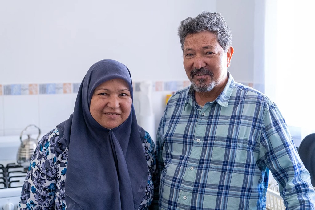 A woman wearing a hijab is smiling and standing next to her husband in their kitchen.